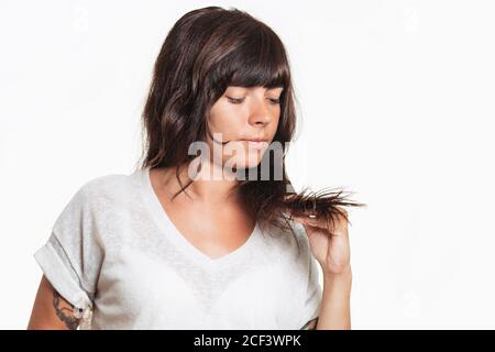 Portrait of a woman with a tattoo on her arm examines the tips of her hair. Copy space. White background. Hair care concept. Stock Photo