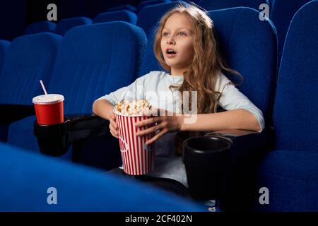 Side view of excited female teen watching action film in cinema. Little girl holding popcorn and sweet water, having rest and relaxing during weekend. Concept of childhood, entertainment. Stock Photo