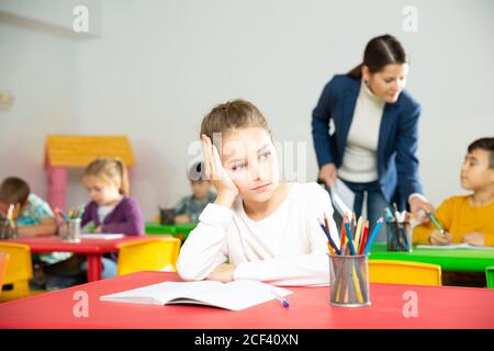 Upset small girl sitting at table in schoolroom on background with pupils studying with teacher Stock Photo