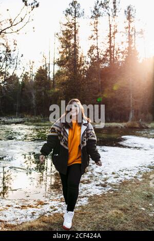 Young joyful female hiker on shore of small pond in snow among trees in sunny woods Stock Photo