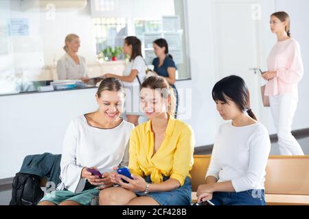 Female students talking and using smartphones, spending time together during recess between lectures in hall Stock Photo