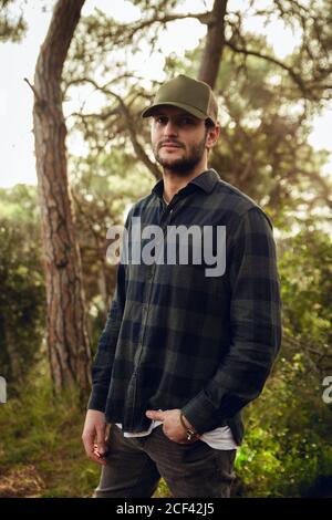 Portrait of handsome brunet man in checkered shirt and baseball cap standing in nature background looking at camera Stock Photo