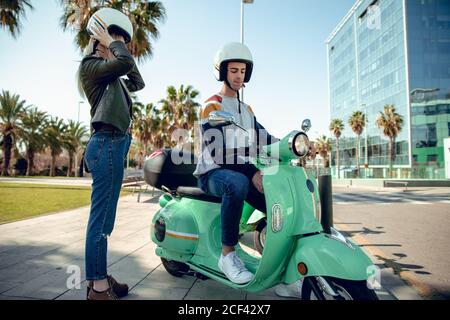 Side view of handsome man in helmet sitting and starting green scooter and young Woman in black jacket wearing helmet Stock Photo