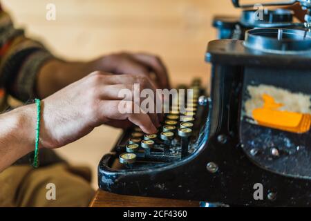 Crop side view of hands of anonymous male in sweater typing on retro typewriter on wooden table at home on Faroe Islands Stock Photo