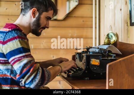 Side view of handsome bearded male in sweater typing on retro typewriter on wooden table at home on Faroe Islands Stock Photo