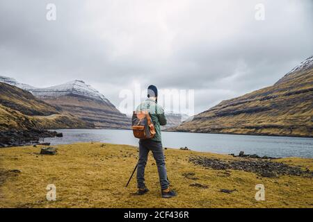 Back view of anonymous bearded man in winter clothes using a photo camera on a tripod outdoors in Faroe Islands landscape Stock Photo