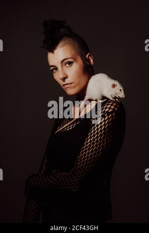 Confident Woman with mohawk carrying white rat on shoulder and looking at camera against black background Stock Photo