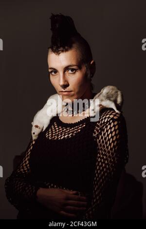 Confident Woman with mohawk carrying white rat on shoulder and looking at camera against black background Stock Photo