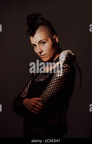 Confident Woman with mohawk carrying white rat on shoulder and looking at camera against black background Stock Photo