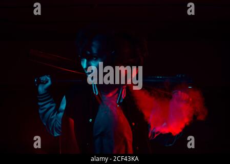Young extraordinary man in bomber jacket with black bat on shoulder in red and blue light on black background Stock Photo