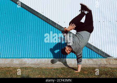 Attractive guy performing handstand and looking at camera while dancing near wall of modern building on city street Stock Photo