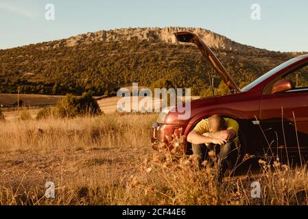 Unhappy adult man embracing knees and crying while sitting on ground near broken car during trip in countryside on sunny day Stock Photo