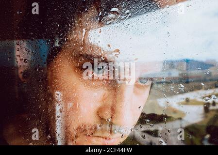 Closeup of sad desperate young man looking away through wet window while staying in self isolation at home because of coronavirus Stock Photo