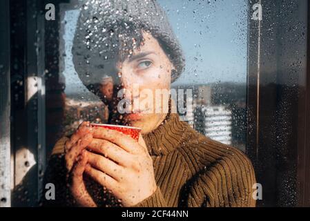 Young depressed female in warm sweater and knitted hat drinking coffee and looking through wet window at city while staying at home during coronavirus pandemic Stock Photo