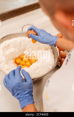 From above unrecognizable confectioner in latex gloves breaking fresh chicken eggs into bowl with wheat flour while preparing pastry in kitchen Stock Photo
