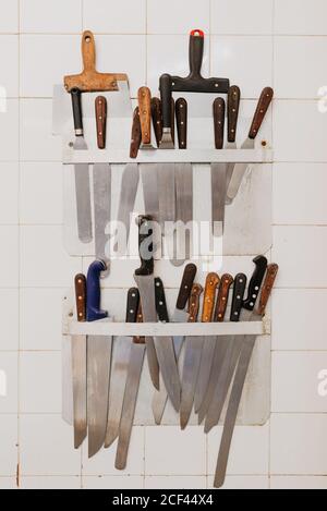 Holders with various knives and spatulas hanging on tiled wall in bakery Stock Photo