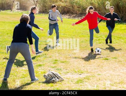 Happy teenage girls and guys playing football on green lawn in spring city park Stock Photo