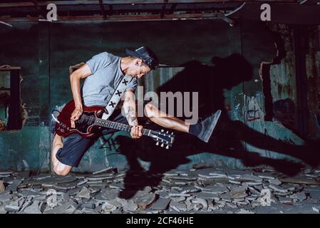 Male musician jumping expressively while playing electric guitar in dark grungy ruined hall Stock Photo