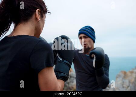 man and Woman in boxing gloves punching each other while standing on cliff against sea and cloudless sky Stock Photo