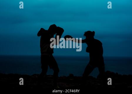 Silhouettes of anonymous fighters practicing kickboxing against sea and dark sky in evening in countryside Stock Photo