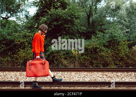 Side view of stylish fearless Woman with bright red suitcase stepping on railroad ties Stock Photo