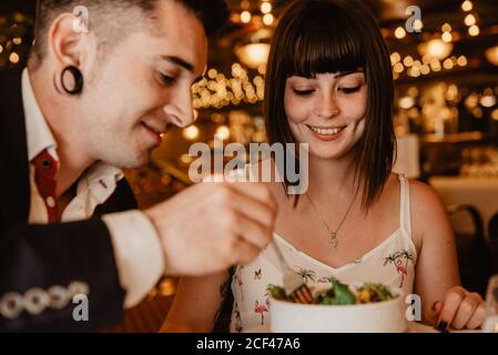 Cheerful young man and Woman eating tasty fresh salad while sitting at table on blurred background of luxury restaurant during romantic dinner Stock Photo