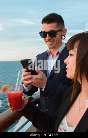 Young handsome man in black sunglasses using mobile phone standing on ship deck while Woman with glass of red drink in sunny evening Stock Photo