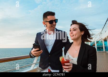 Young handsome man in black sunglasses using mobile phone standing on ship deck while Woman with glass of red drink in sunny evening Stock Photo