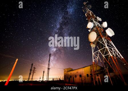 From below wonderful view of media antenna and silhouettes of electricity high voltage power towers against night city on horizon and gorgeous Milky Way in clear sky