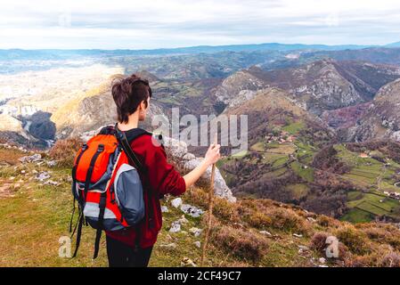 Back view of unrecognizable hiker with backpack and stick standing with arms outstretched and enjoying freedom viewing majestic scenery of countryside located along river shore in valley against foggy ridges at horizon under cloudy sky in Spain Stock Photo