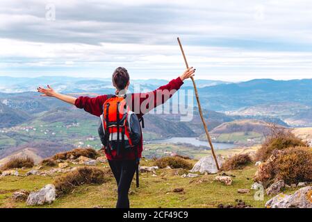 Back view of unrecognizable hiker with backpack and stick standing with arms outstretched and enjoying freedom viewing majestic scenery of countryside located along river shore in valley against foggy ridges at horizon under cloudy sky in Spain Stock Photo