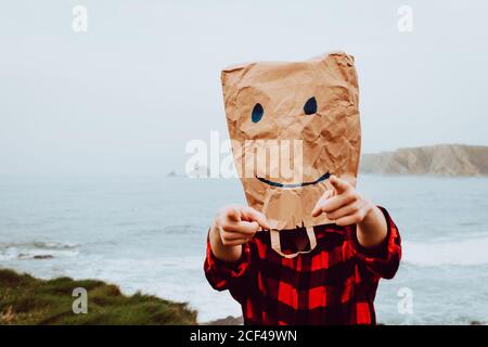 unrecognizable person wearing paper bag with drawn happy grimace on head and pointing at camera while standing at seaside Stock Photo