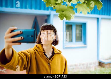 Young Woman in yellow warm jacket smirking and using smartphone to take selfie on blurred background of countryside cottage Stock Photo