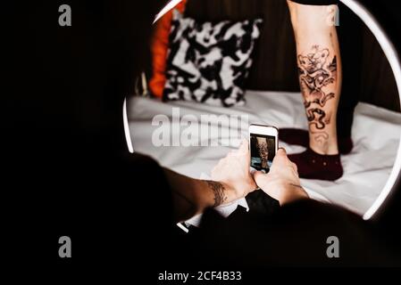 unrecognizable man tattooer using smartphone to take picture of tattoo on leg of crop customer for portfolio in contemporary studio Stock Photo