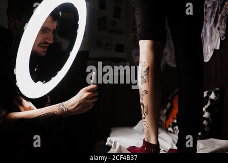 unrecognizable man tattooer using smartphone to take picture of tattoo on leg of crop customer for portfolio in contemporary studio Stock Photo