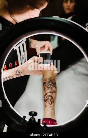unrecognizable man tattooer using smartphone to take picture of tattoo on leg of crop customer for portfolio in contemporary studio Stock Photo