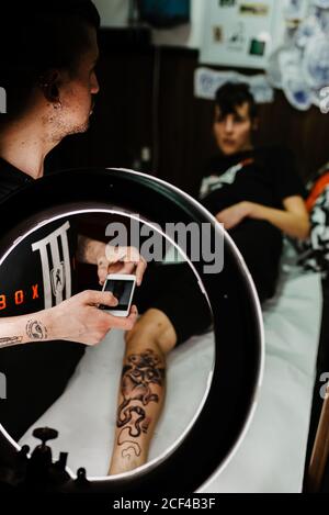 unrecognizable man tattooer using smartphone to take picture of tattoo on leg of crop customer for portfolio in contemporary studio Stock Photo