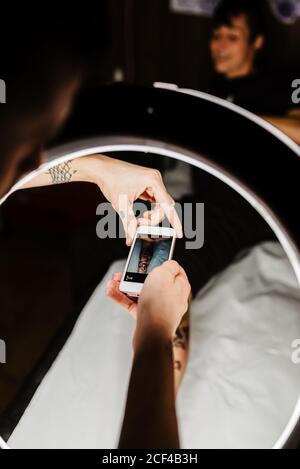 unrecognizable man tattooer using smartphone to take picture of tattoo on leg of crop customer for portfolio in contemporary studio Stock Photo
