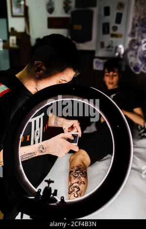 unrecognizable man tattooer using smartphone to take picture of tattoo on leg of crop customer for portfolio in contemporary studio Stock Photo