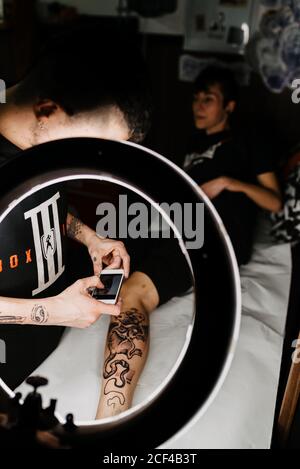 unrecognizable man tattooer using smartphone to take picture of tattoo on leg of crop customer for portfolio in contemporary studio Stock Photo