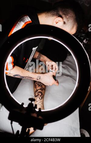 unrecognizable man tattooer using smartphone to take picture of tattoo on leg of crop customer for portfolio in contemporary studio Stock Photo