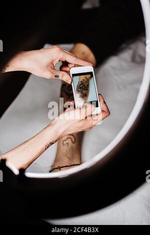 unrecognizable man tattooer using smartphone to take picture of tattoo on leg of crop customer for portfolio in contemporary studio Stock Photo