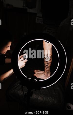 unrecognizable man tattooer using smartphone to take picture of tattoo on leg of crop customer for portfolio in contemporary studio Stock Photo