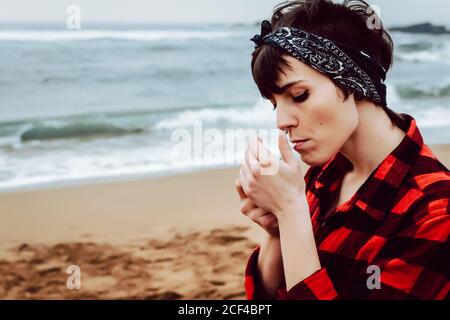 Side view of young informal female wearing casual checkered shirt and headband lighting cigarette while standing on sandy beach with stormy sea in background Stock Photo