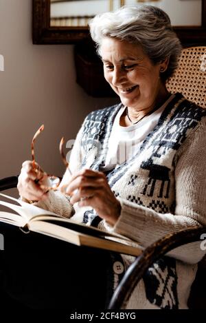 Relaxed mature Woman with gray hair and kind smile reading book holding glasses in hand near window in armchair Stock Photo