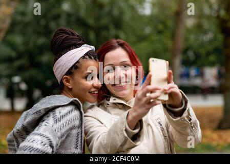 Lovely multiethnic couple of smiling women staying close and taking selfie on mobile phone in autumn park Stock Photo