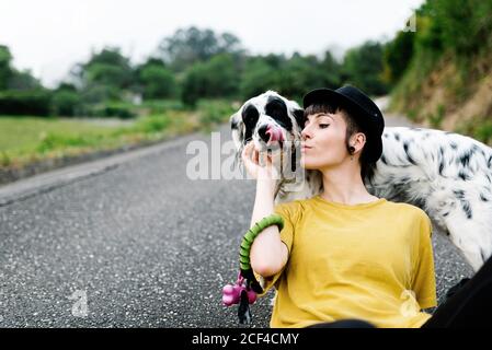 Positive young lady in casual clothes and black hat resting on ground with dog during walk in park Stock Photo