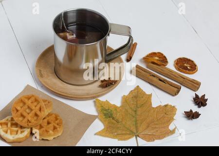 Mug of tea and waffles. Nearby are dried maple leaves, cinnamon, anise and dried oranges on the table. Autumn still life. Stock Photo