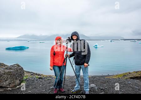 Blue glacier icebergs in lagoon lake in Iceland with photographer photography couple man woman landscape view on cloudy day with ice floating on water Stock Photo