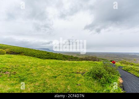 Skaftafell, Iceland green summer landscape and man in poncho walking hiking on steep trail path hiking road covered and cloudy sky Stock Photo